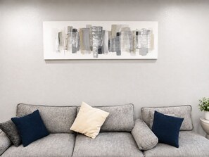 A grey sofa with two dark blue pillows and a beige pillow, a white abstract painting on the wall, and a potted plant on a side table.