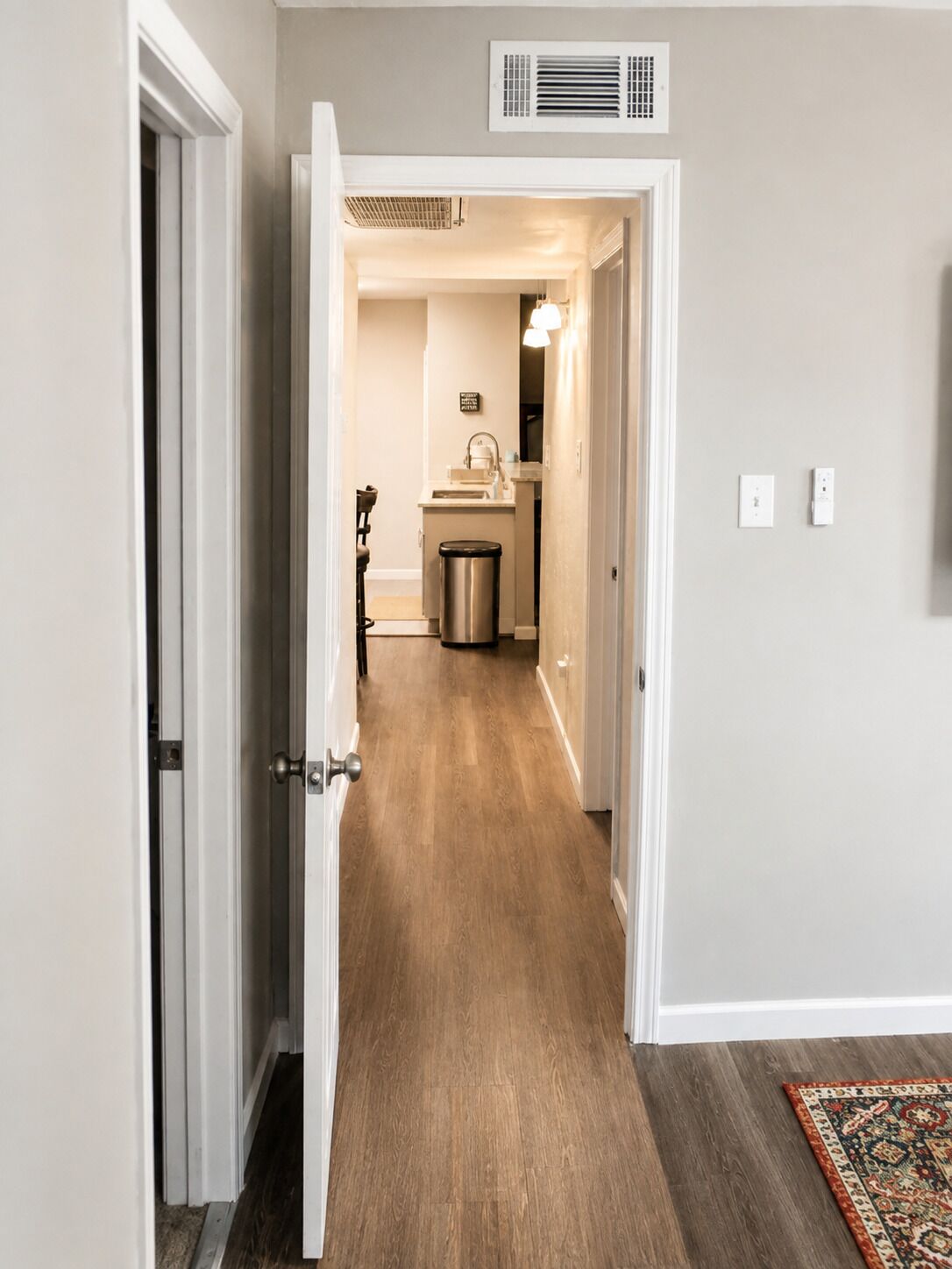 A hallway with a wooden floor leading to a bathroom with a sink and a trash can.