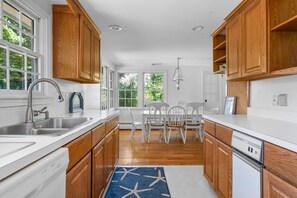 Una cocina con armarios de madera, encimera blanca, un fregadero y una zona de comedor con sillas blancas.