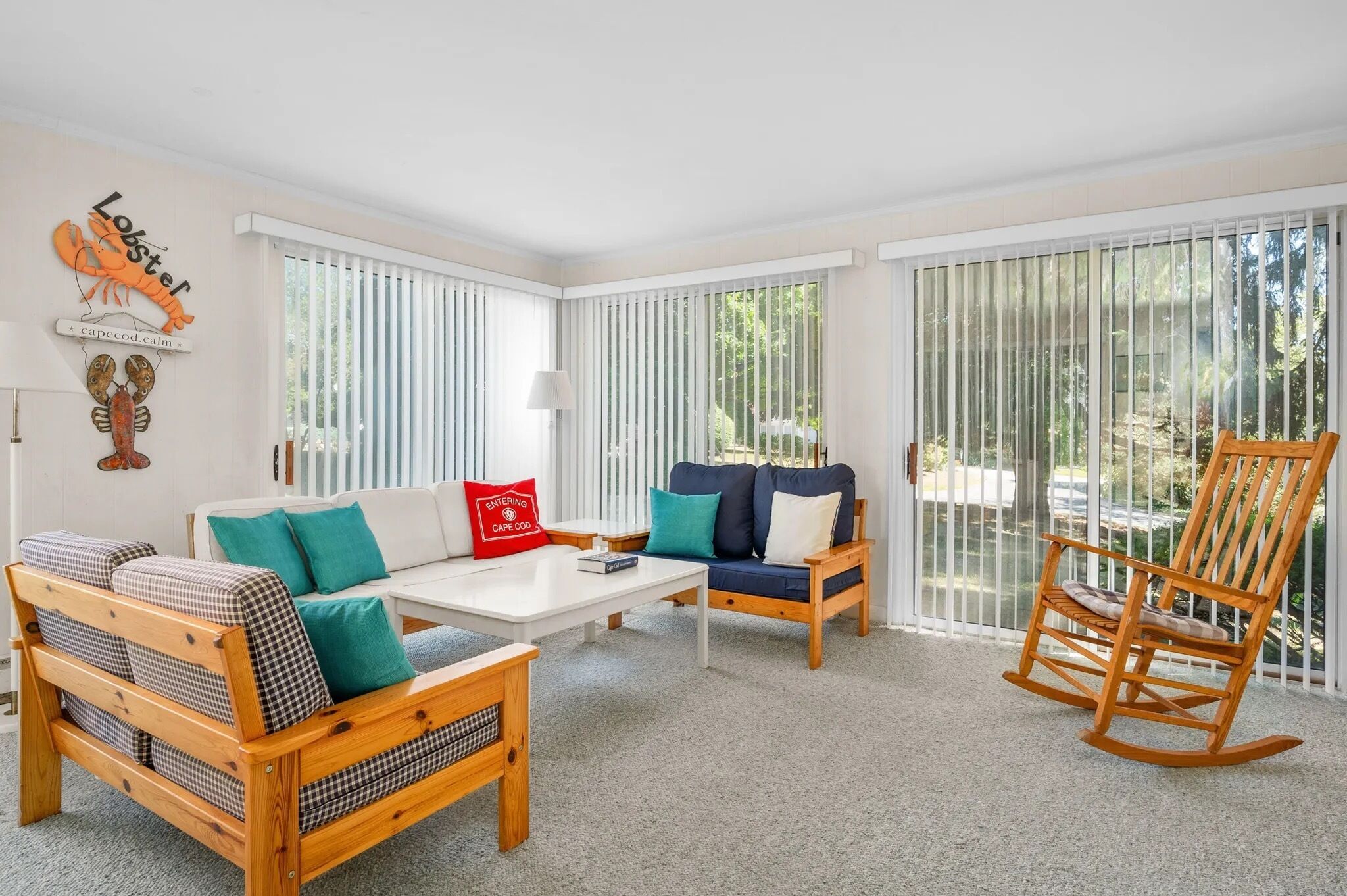 A room with a white ceiling, a wooden rocking chair, a sofa with teal cushions, a white coffee table, and a red pillow with 'ENTERING CAPE COD' text.