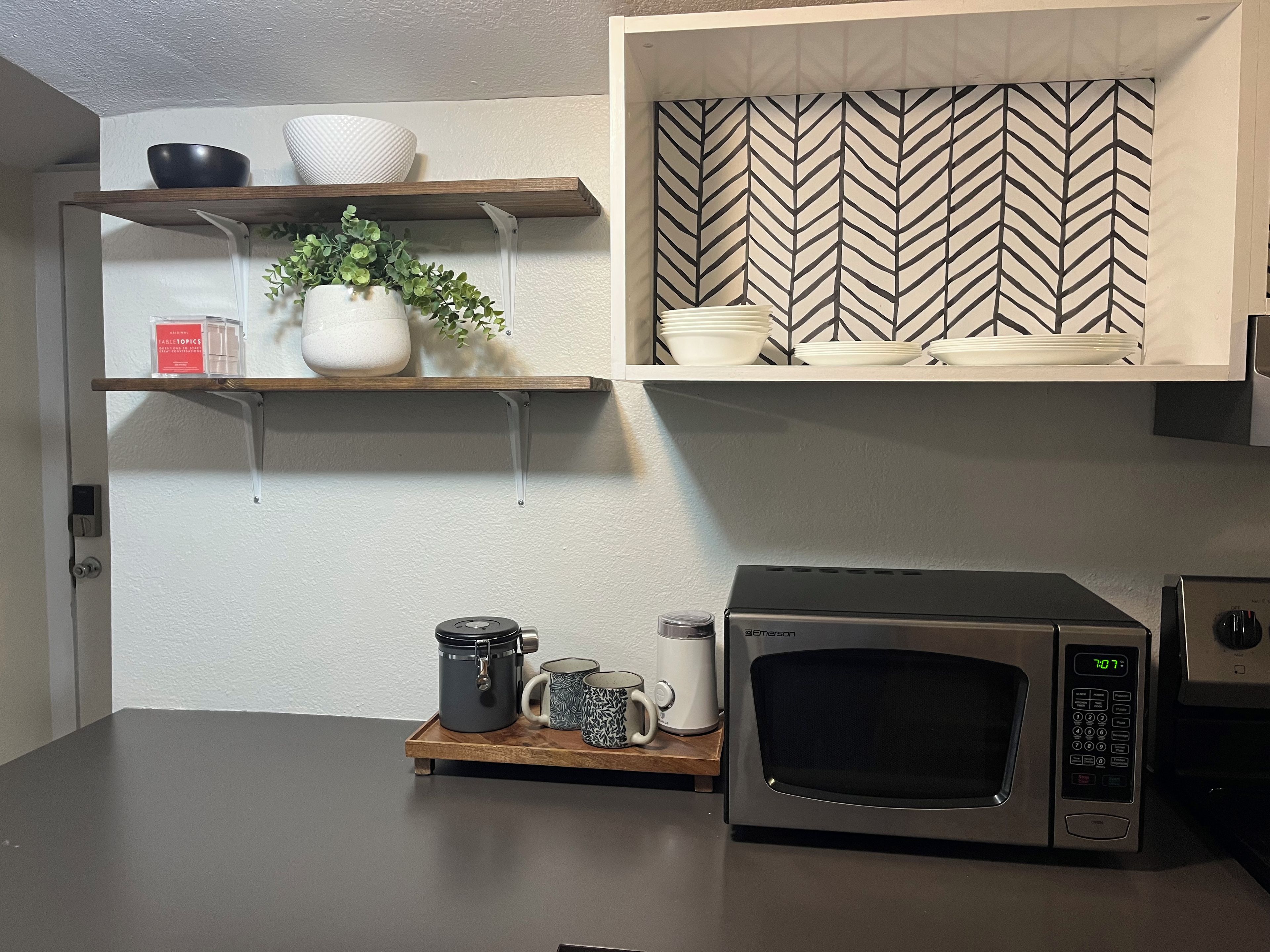 Una cocina con microondas, estante con cuencos y una planta, y un salpicadero con estampado de espiga.