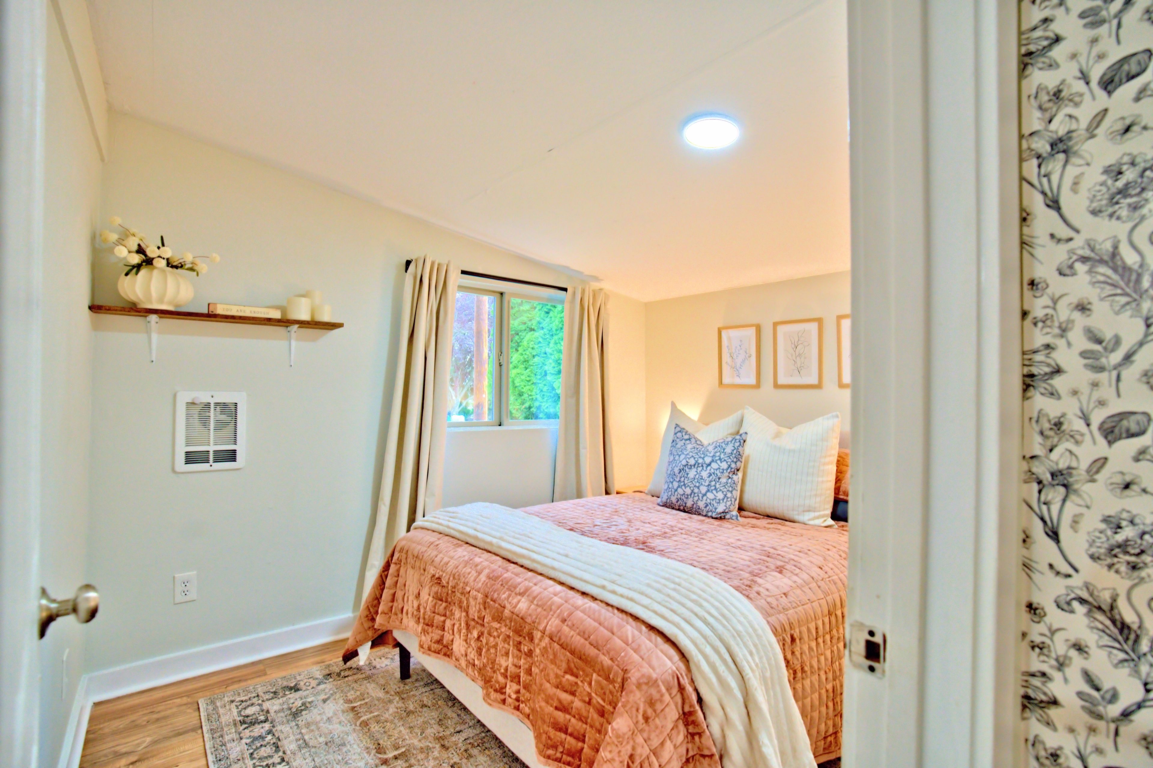 A bedroom with a bed, a window, a radiator, a shelf with decorative items, and a floral-patterned curtain.