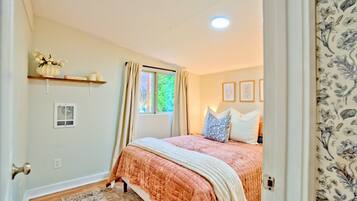 A bedroom with a bed, a window, a radiator, a shelf with decorative items, and a floral-patterned curtain.