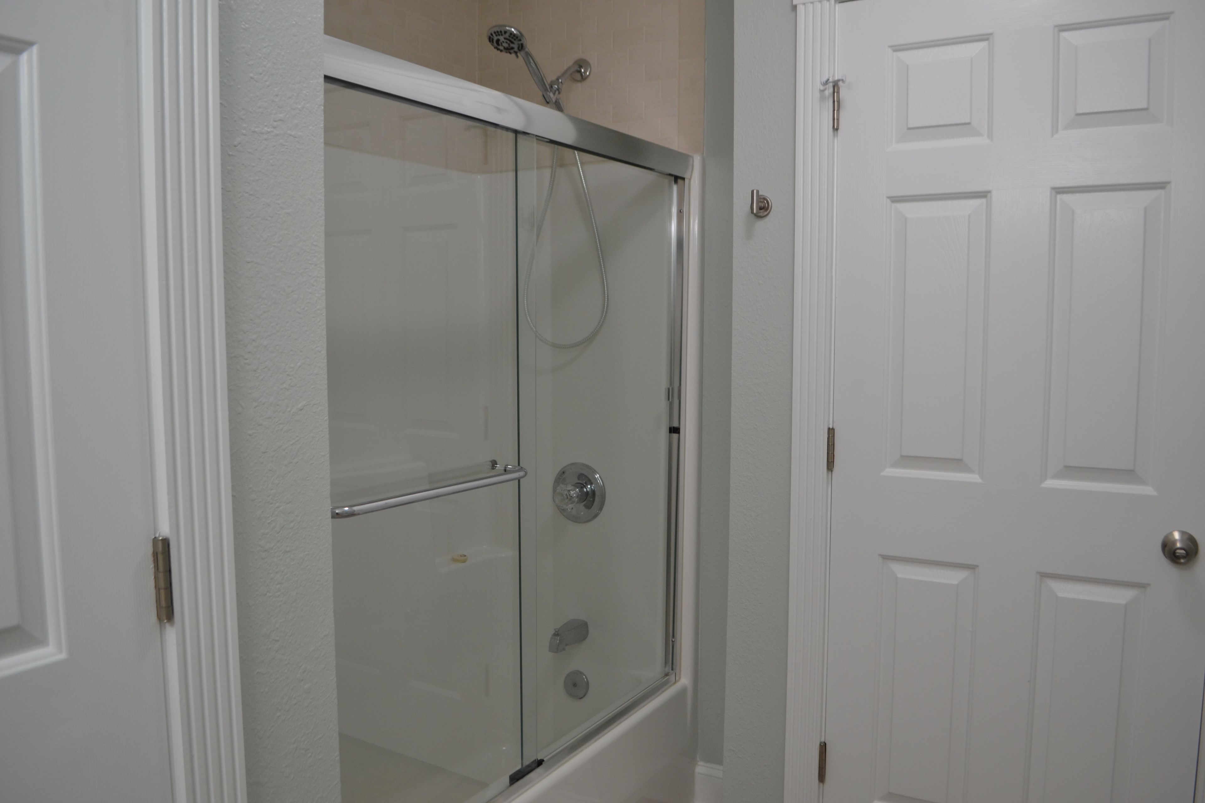 A glass-enclosed shower with a handheld showerhead, a glass door, and a white door with a handle.