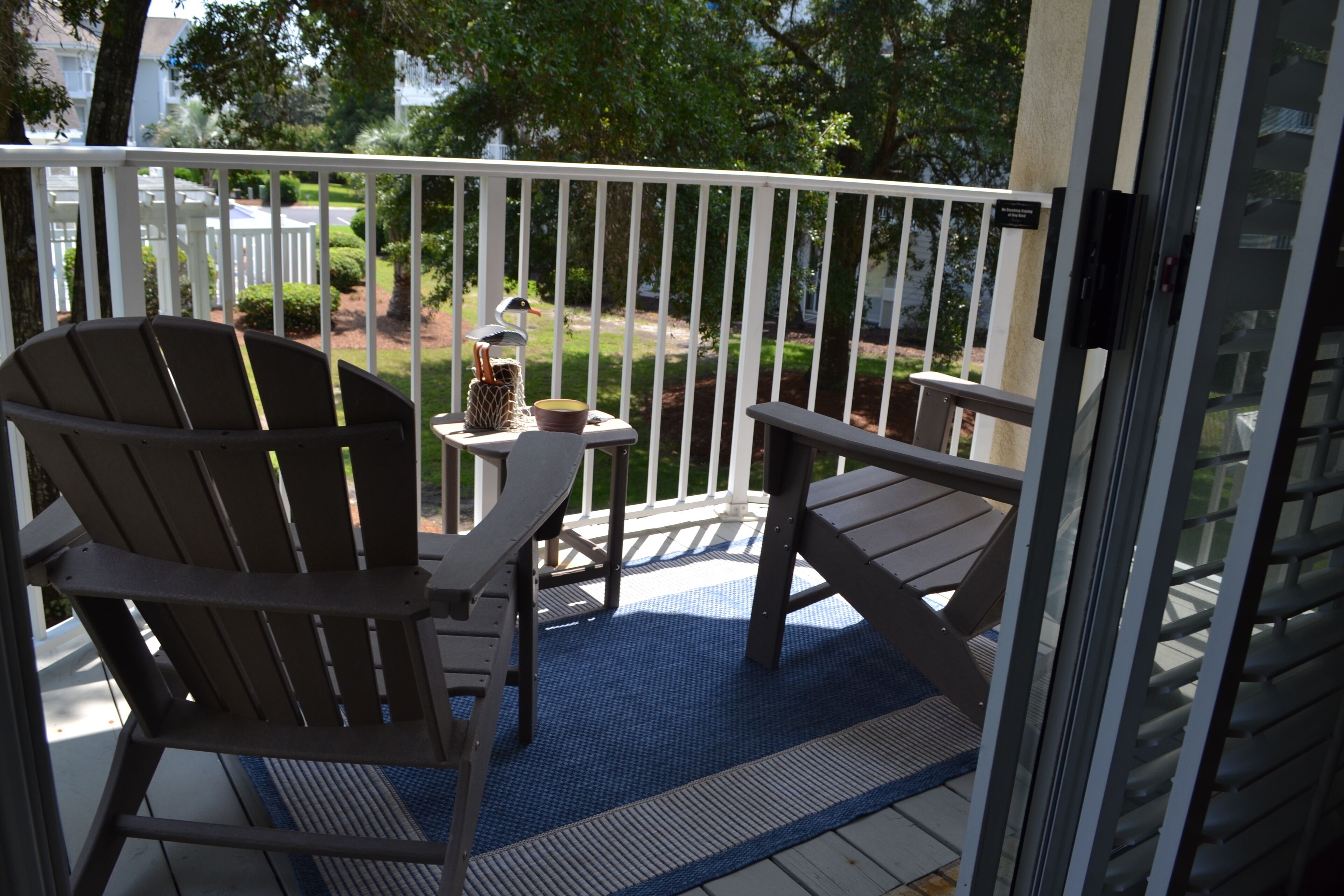 A balcony with two Adirondack chairs, a small table with a birdcage ornament, and a blue mat.