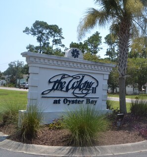 A sign for 'The Colony at Oyster Bay' with a decorative top and surrounded by grass and a palm tree.