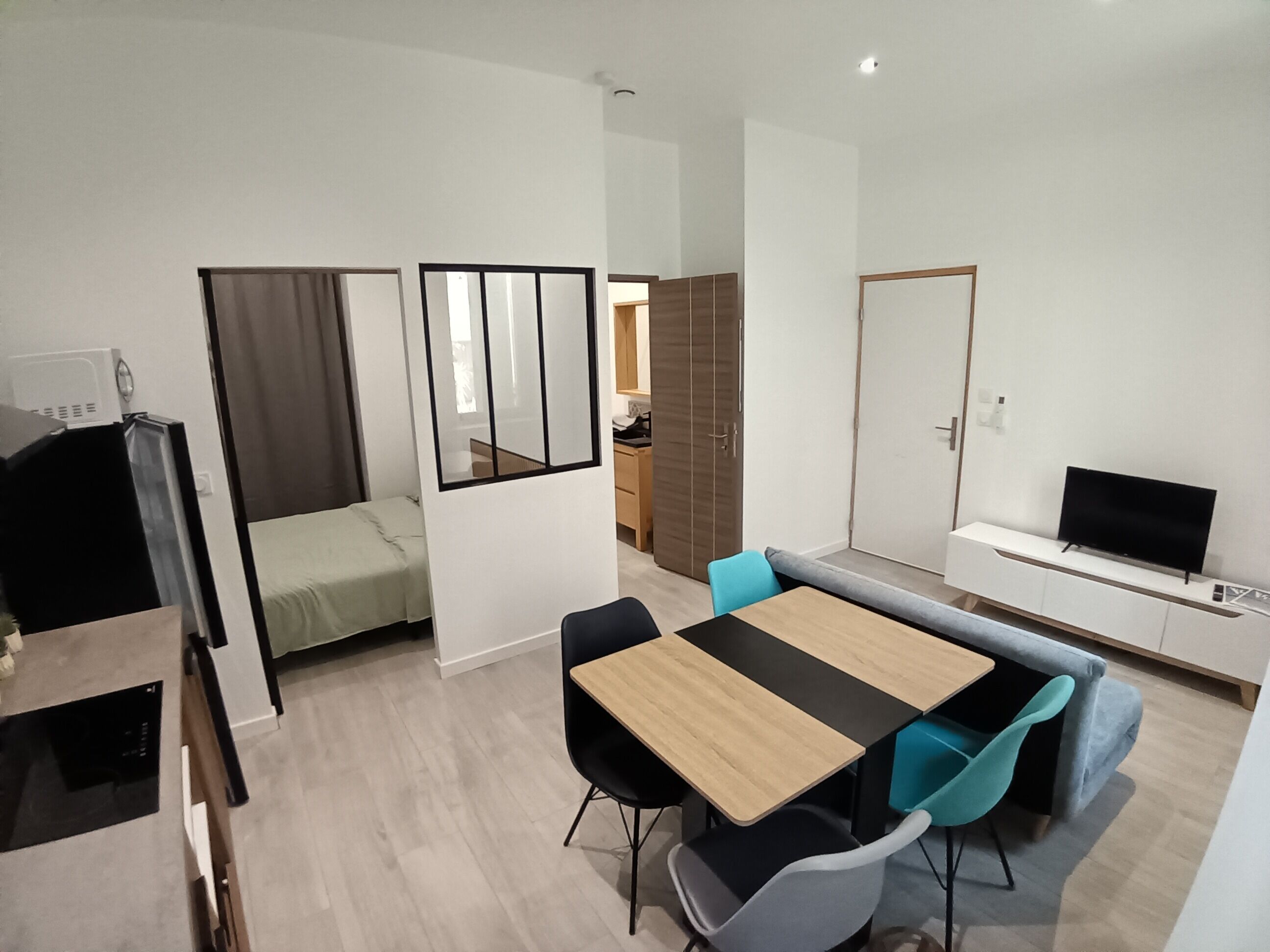 Ein moderner, gut beleuchteter Raum mit Esstisch, Stühlen, Fernseher und einem Bett mit Vorhängen.