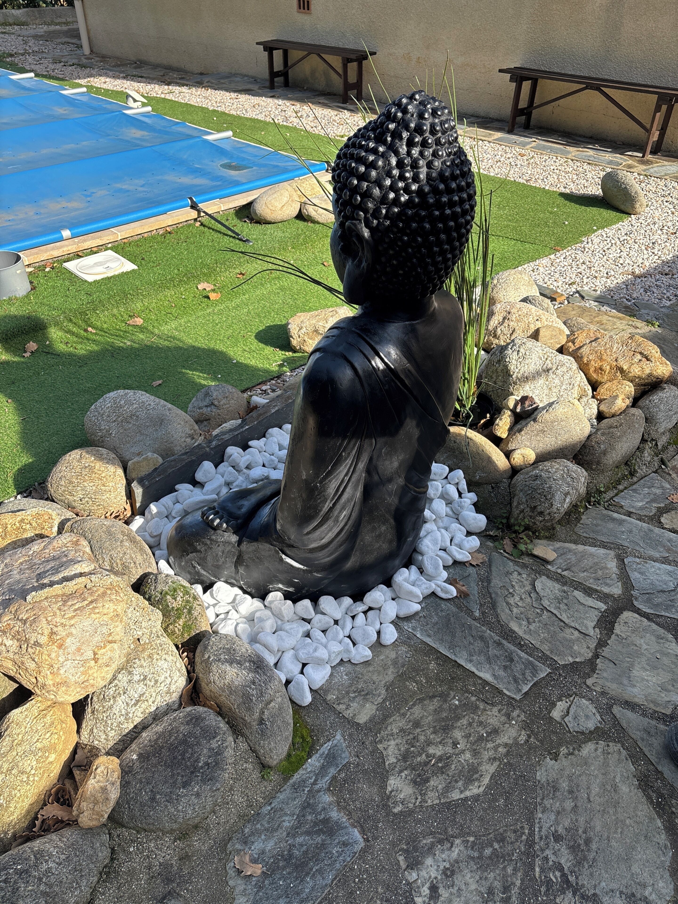 A statue of a seated figure in a garden setting with a pool, rocks, and a bench.