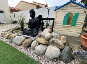 A Buddha statue in a garden setting with a playhouse and a white building in the background. - Charming family home with a private pool (Laroque-des-Albères)