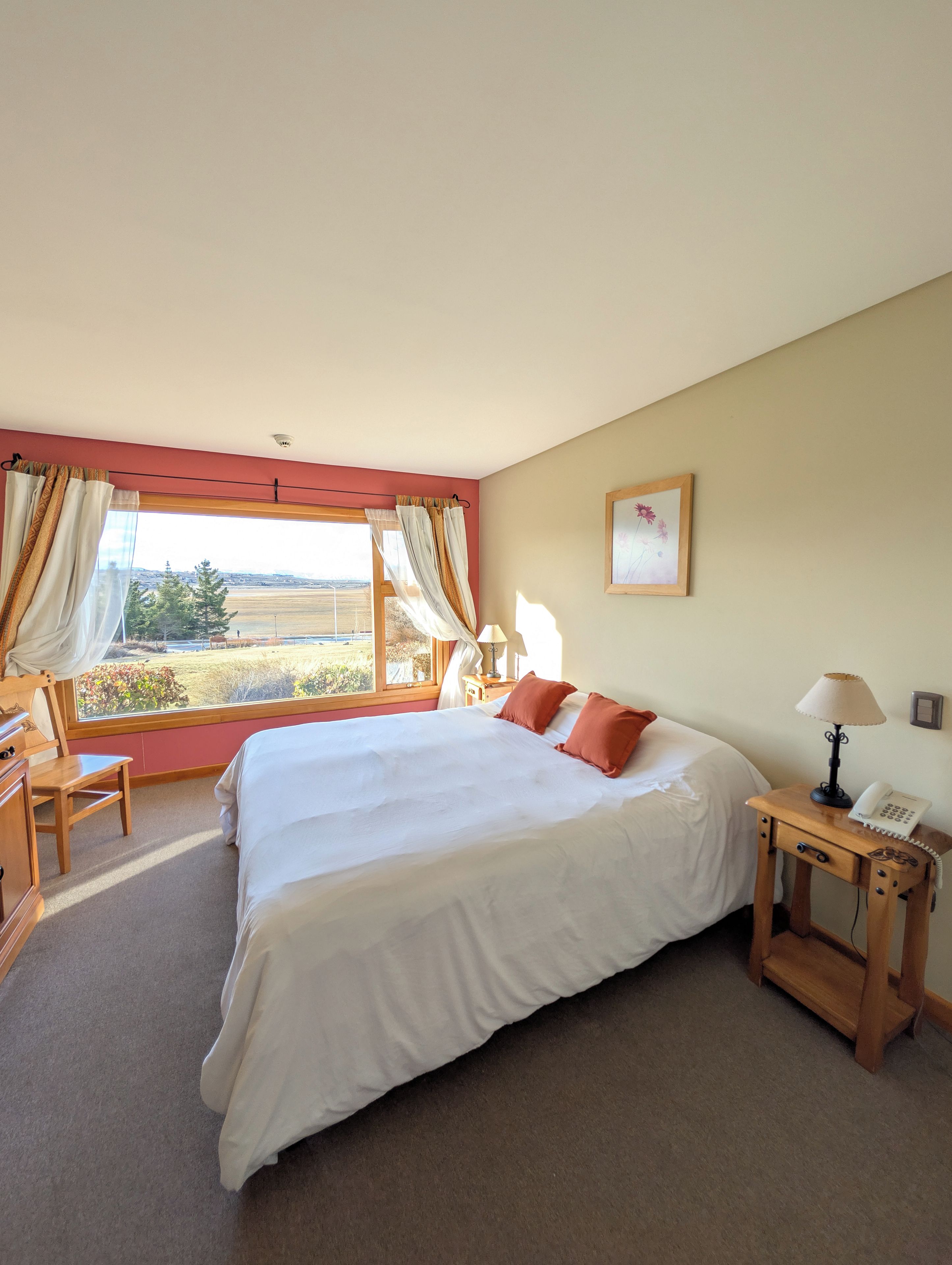 A bedroom with a large bed, a desk, a chair, a nightstand with a lamp, and a view of the countryside.