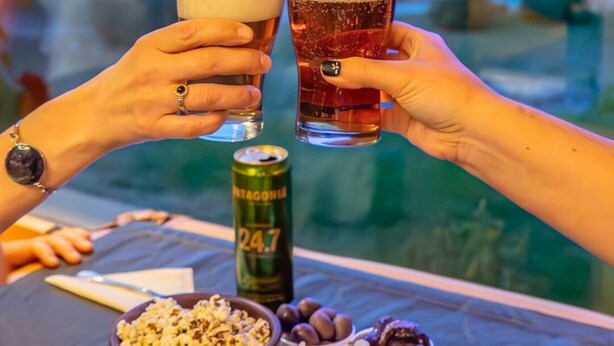 Dos personas brindando con vasos de cerveza, una lata de cerveza y varios bocadillos sobre una mesa con vista al agua.