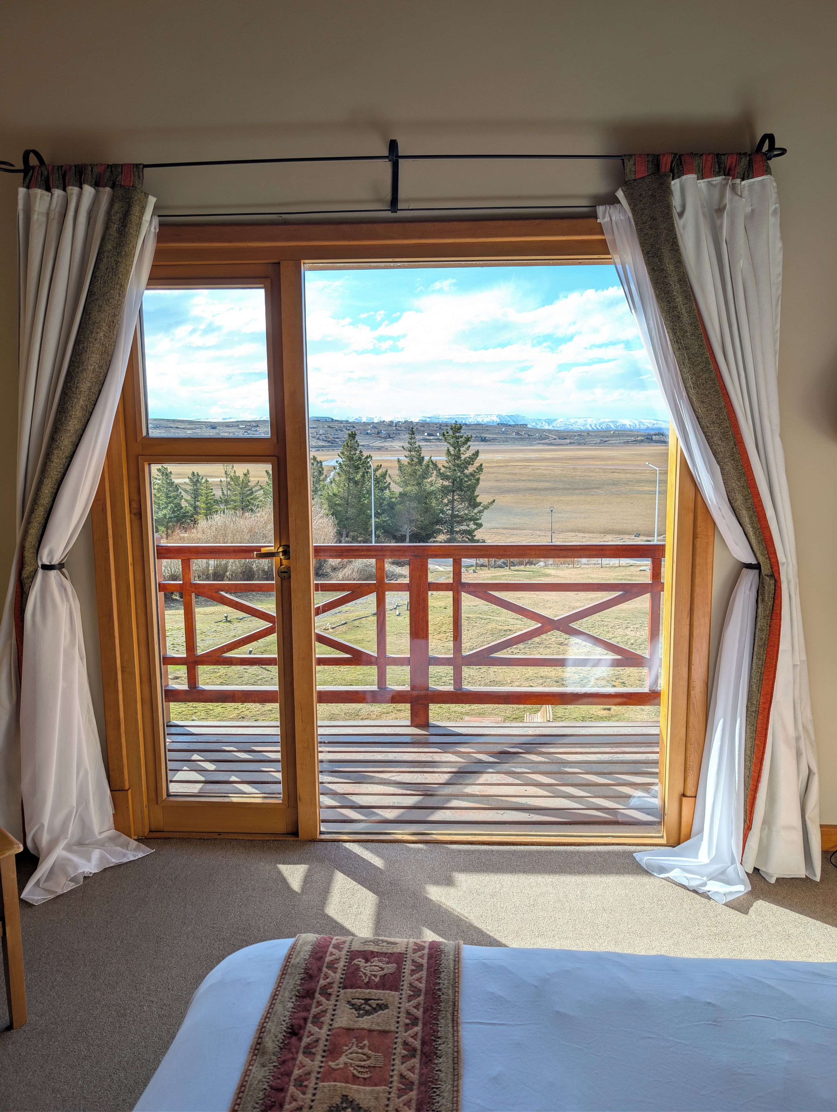Una habitación con una cama, vista a una terraza de madera y un paisaje con árboles y montañas.