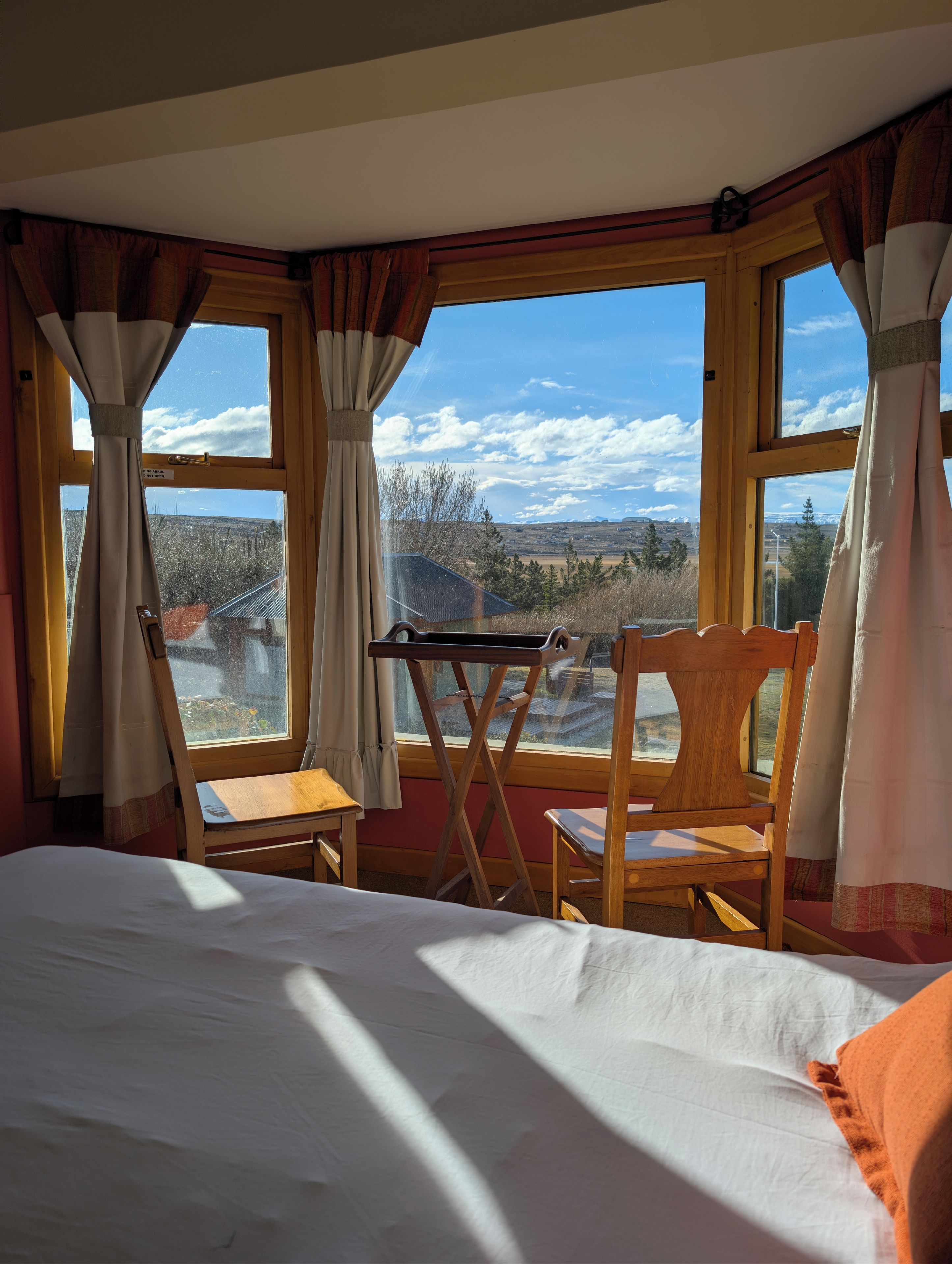A room with a bed, two wooden chairs, a small table, and large windows offering a view of a mountainous landscape.