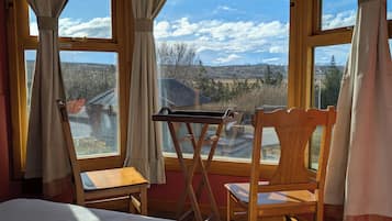 Una habitación con una cama, dos sillas de madera, una mesita y ventanas amplias con vistas a un paisaje montañoso.