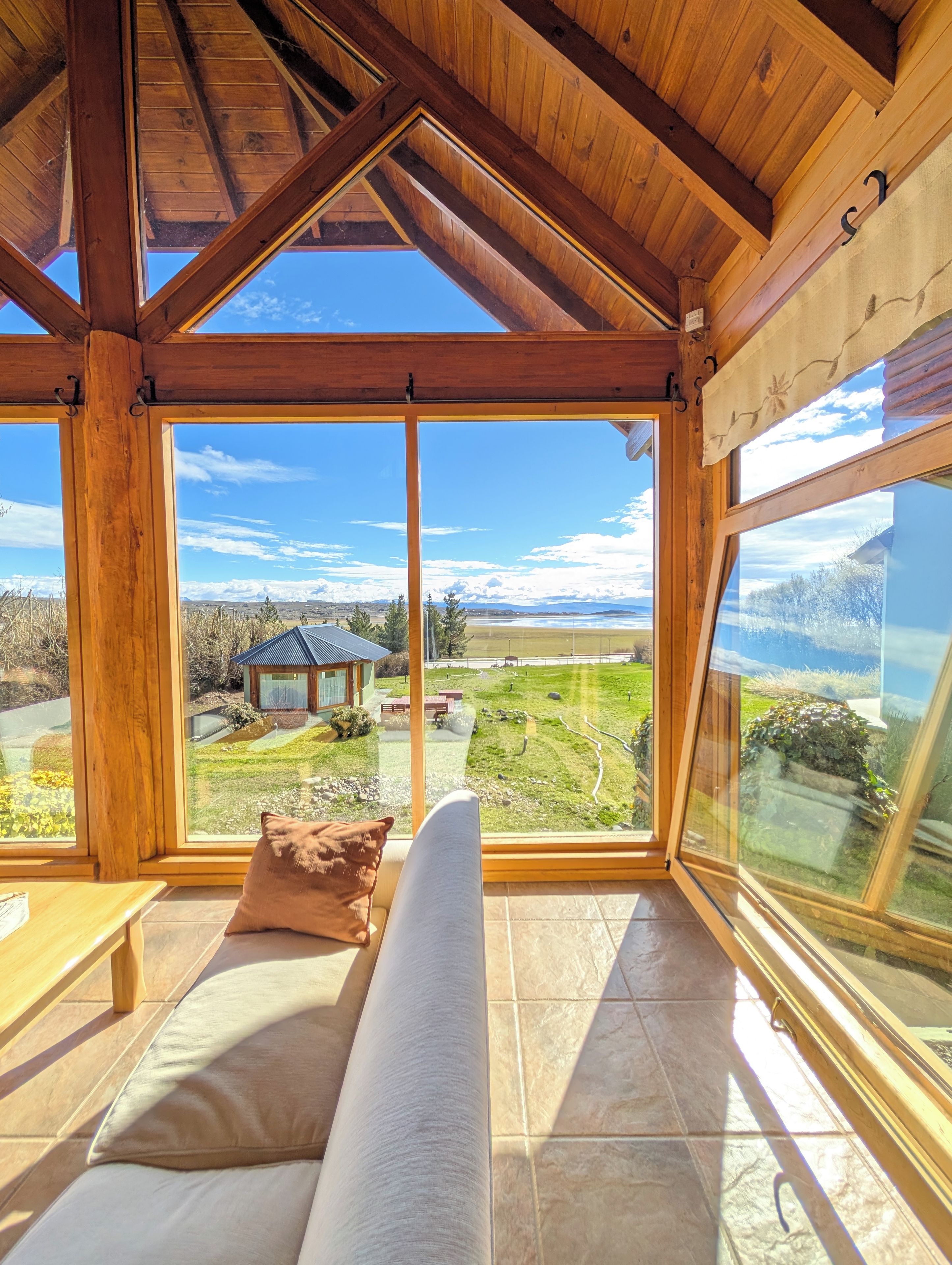 A room with a wooden ceiling, large windows, a bench, and a view of a landscape with a building.