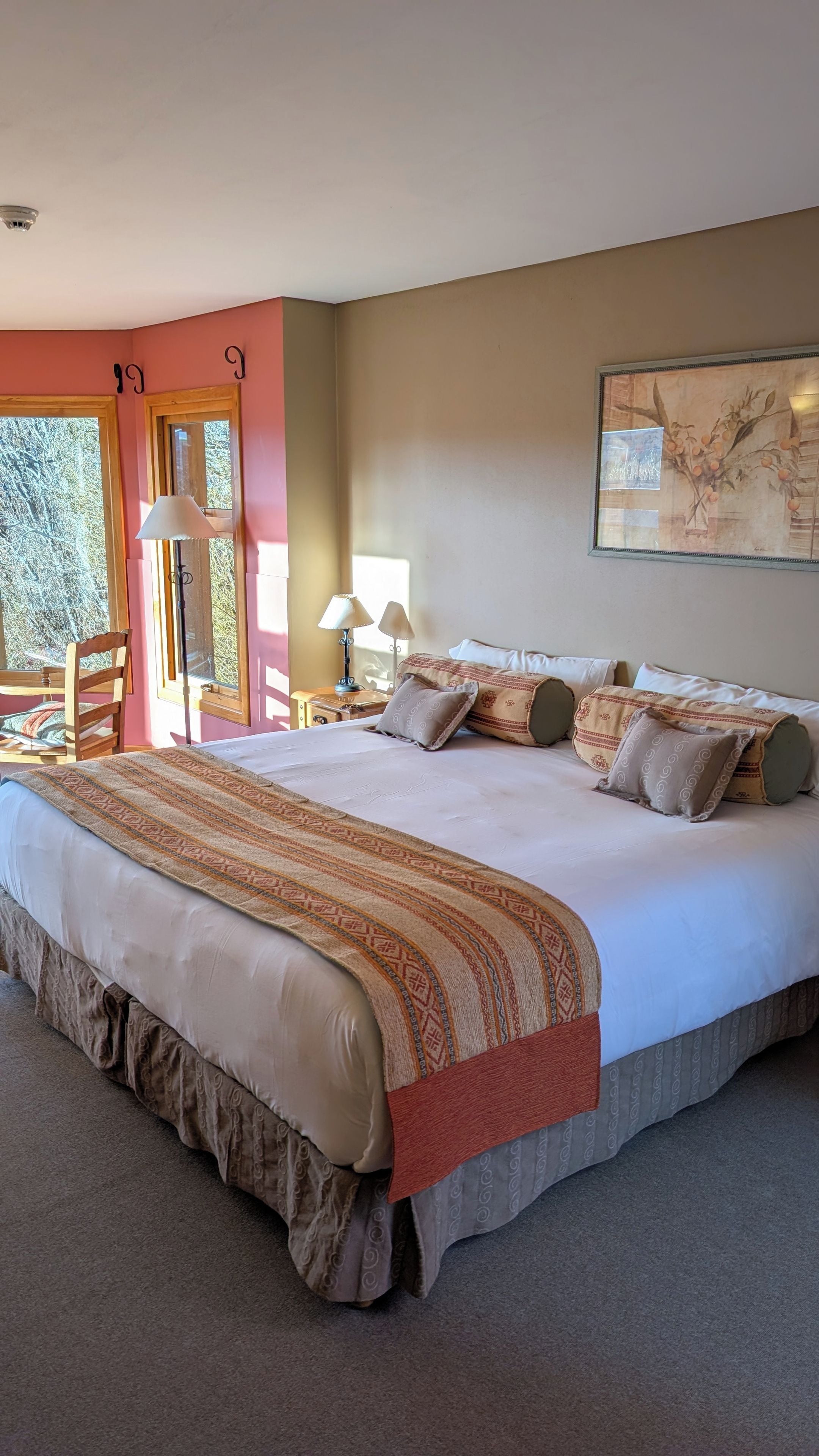 A large bed with multiple pillows and a patterned bedspread, a nightstand with lamps, and a painting on the wall.