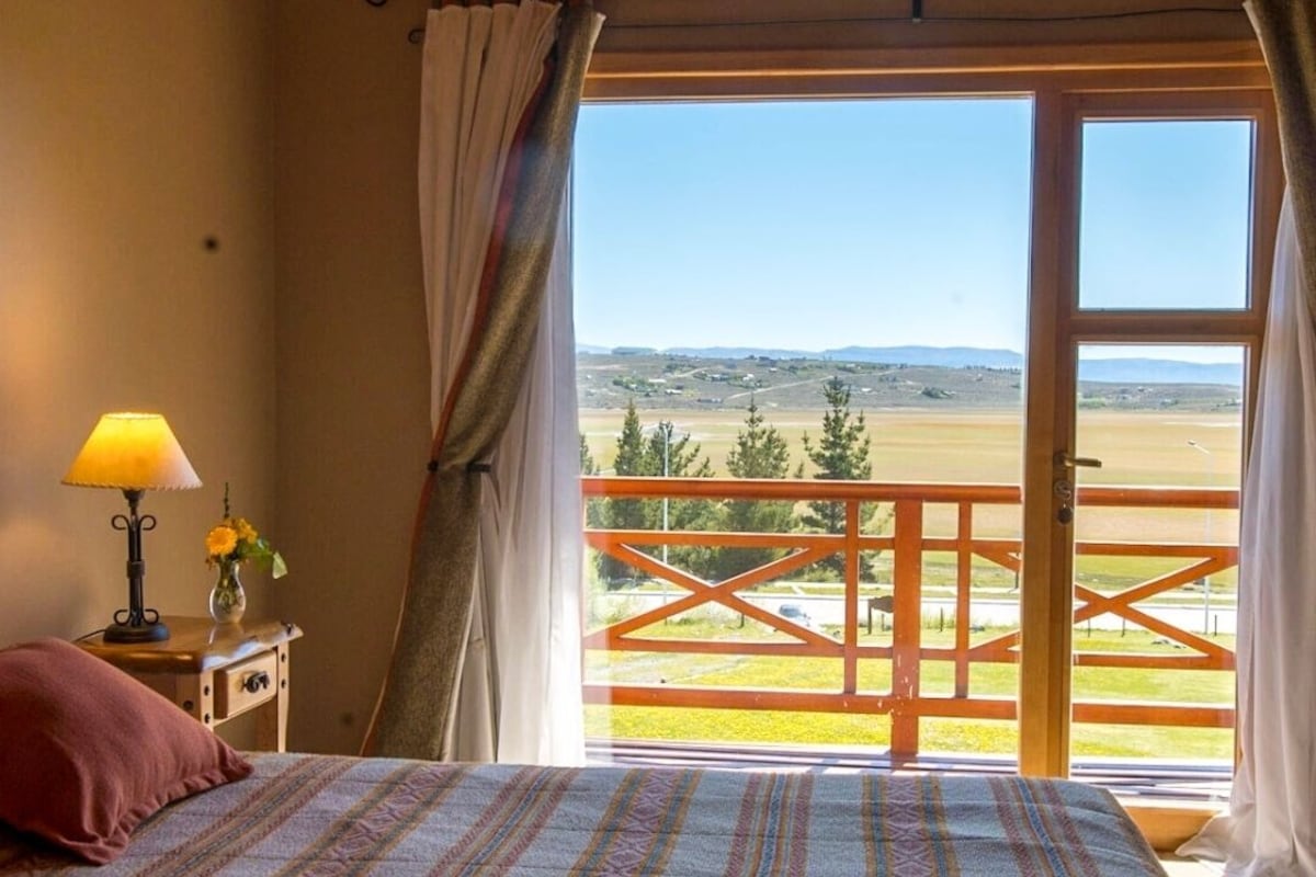 A bedroom with a bed, a nightstand, a lamp, and a view of a balcony with a railing.