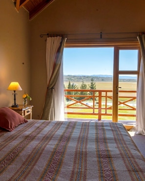 A bedroom with a bed, a nightstand, a lamp, and a view of a balcony with a railing. - Hostería Los Hielos (El Calafate)
