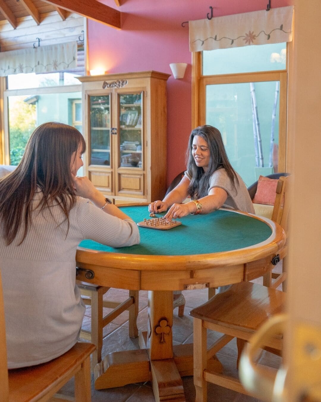 Deux personnes sont assises autour d’une table en bois, recouverte d’un tapis vert, et jouent à un jeu de société. La pièce est équipée d’une armoire et d’une fenêtre.