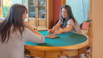 Deux personnes sont assises autour d’une table en bois, recouverte d’un tapis vert, et jouent à un jeu de société. La pièce est équipée d’une armoire et d’une fenêtre.