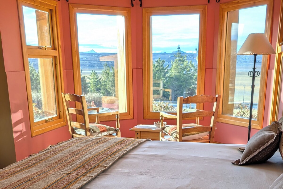 Quarto com cama grande, duas cadeiras, uma mesinha e vista para o exterior.