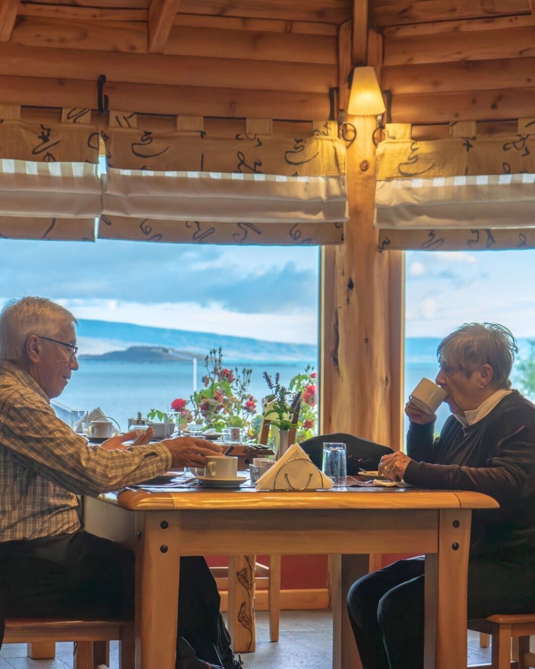 Un interior de madera con una mesa dispuesta para dos, una ventana con vistas al mar y una lámpara colgando del techo.