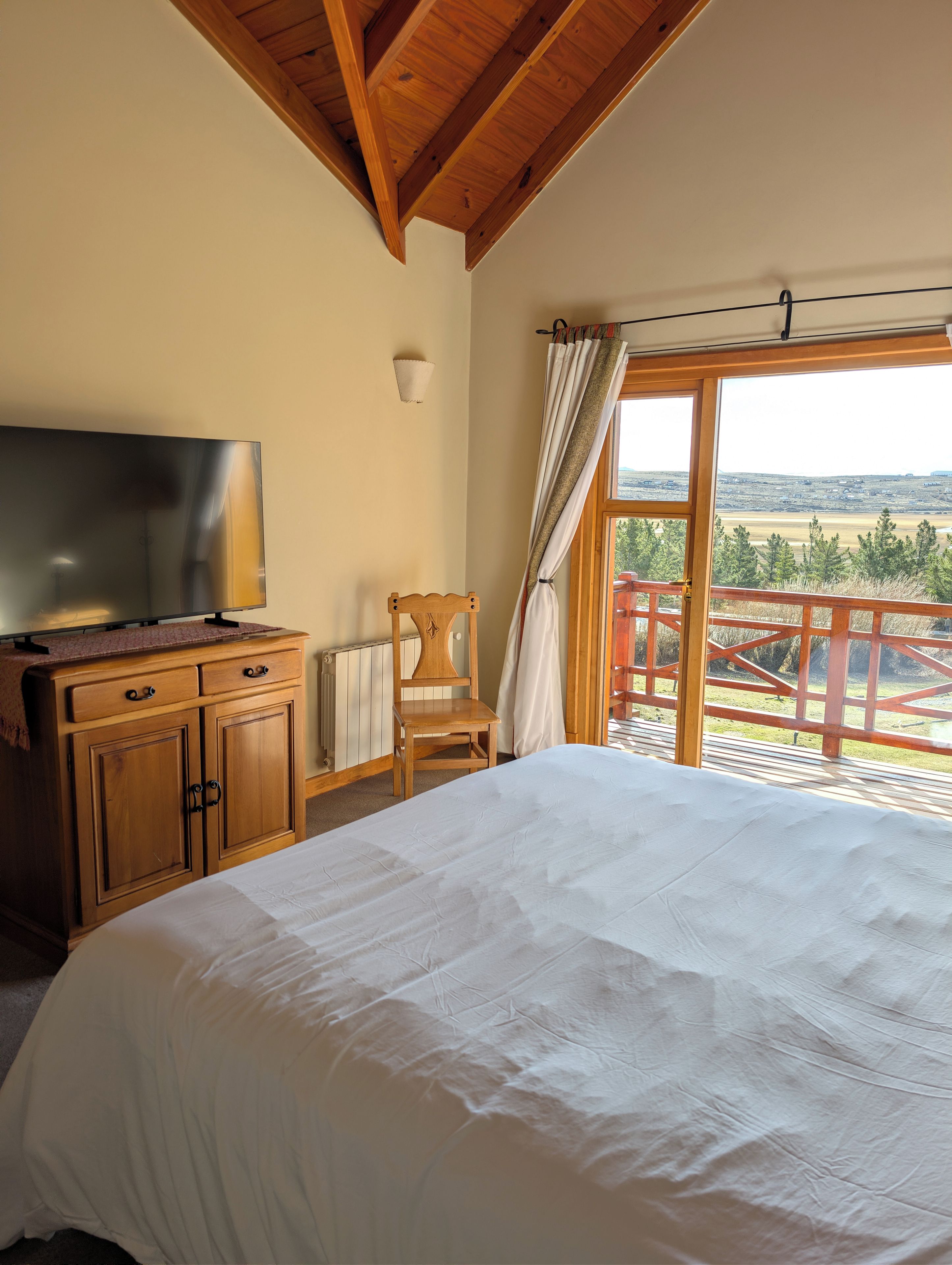 A bedroom with a bed, a TV on a wooden dresser, a chair, and a view of a balcony with a railing.