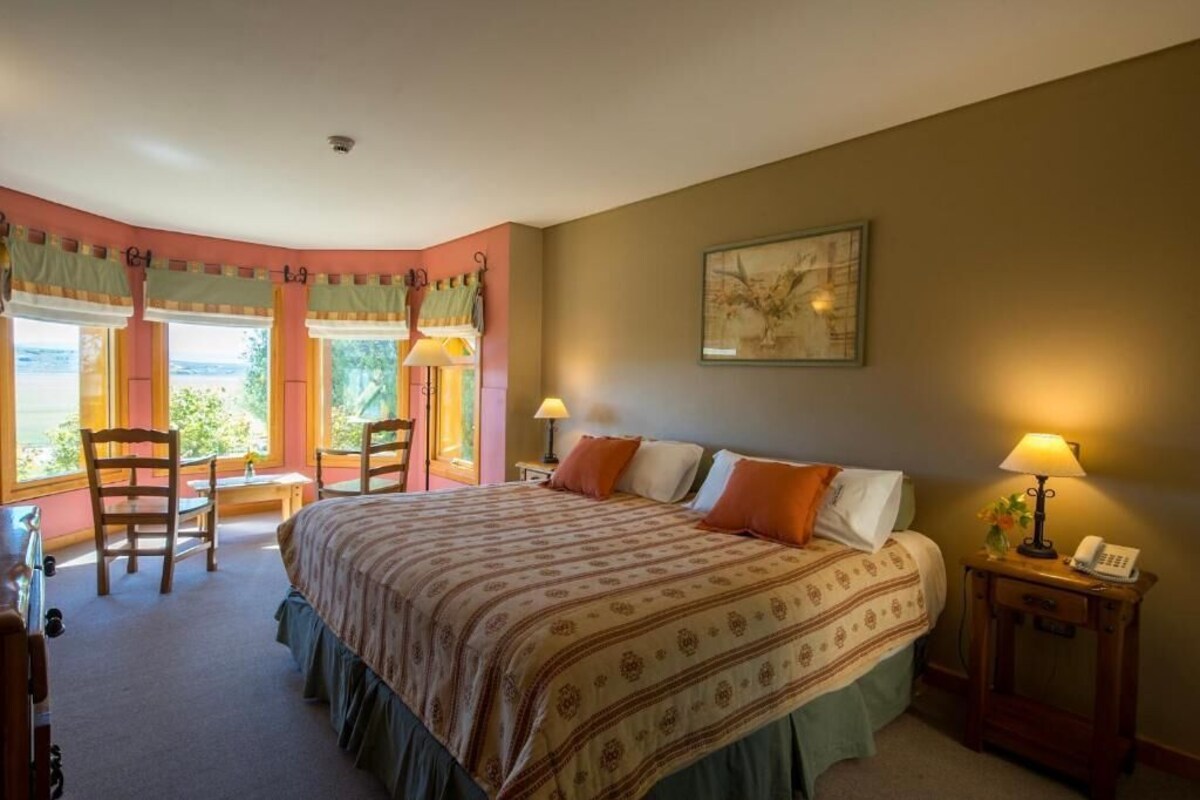A bedroom with a large bed, two bedside lamps, a chair, a desk, and a window with curtains.