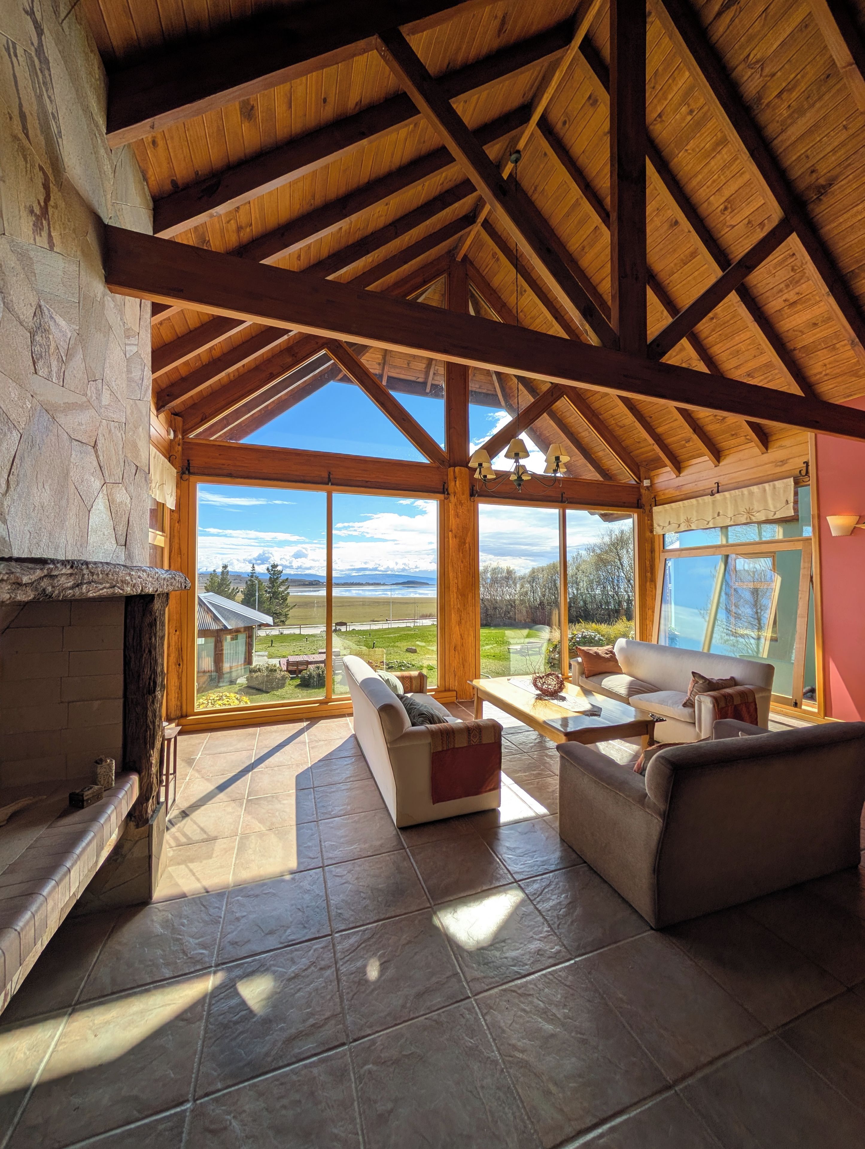Amplia sala de estar con chimenea de piedra, vigas de madera y ventanas grandes que ofrecen vistas al exterior.