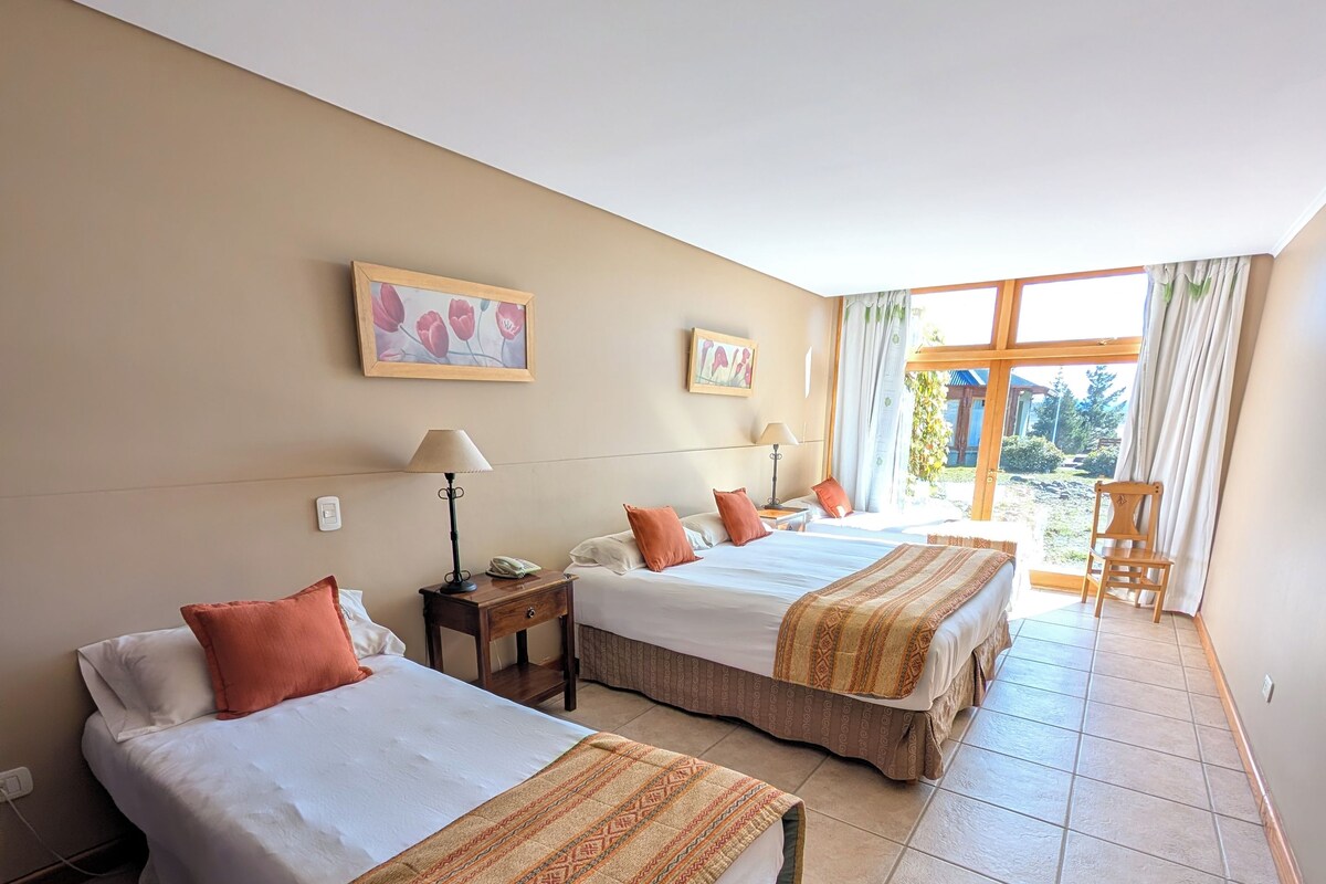 Quarto de hotel com duas camas, uma mesinha, uma luminária e uma janela com cortinas.