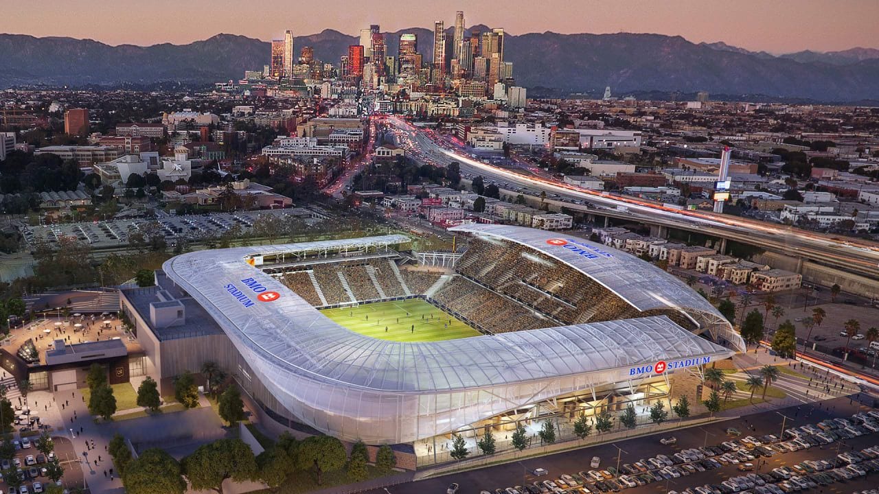 A stadium with a large, transparent roof, surrounded by parking and roads, with a city skyline in the background.