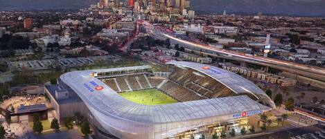 A stadium with a large, transparent roof, surrounded by parking and roads, with a city skyline in the background.