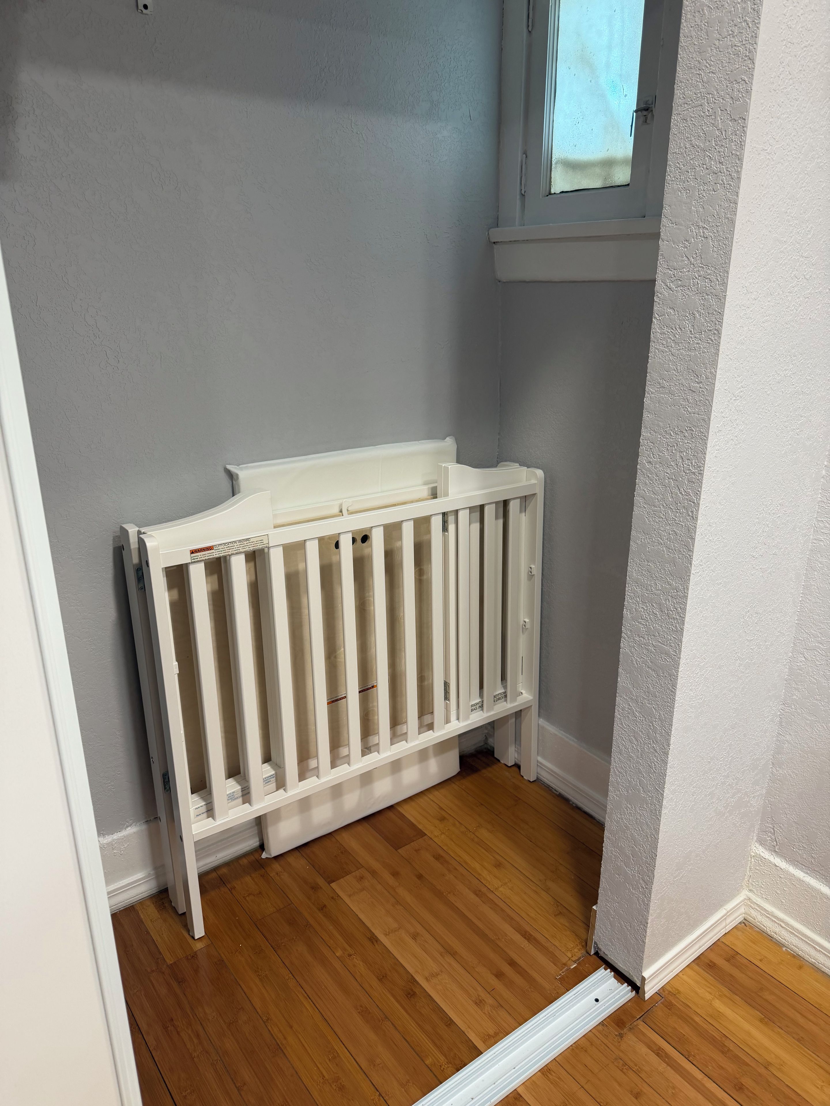 A corner of a room with a white crib against a grey wall, wooden flooring, and a window with a view.