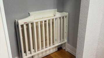 A corner of a room with a white crib against a grey wall, wooden flooring, and a window with a view.