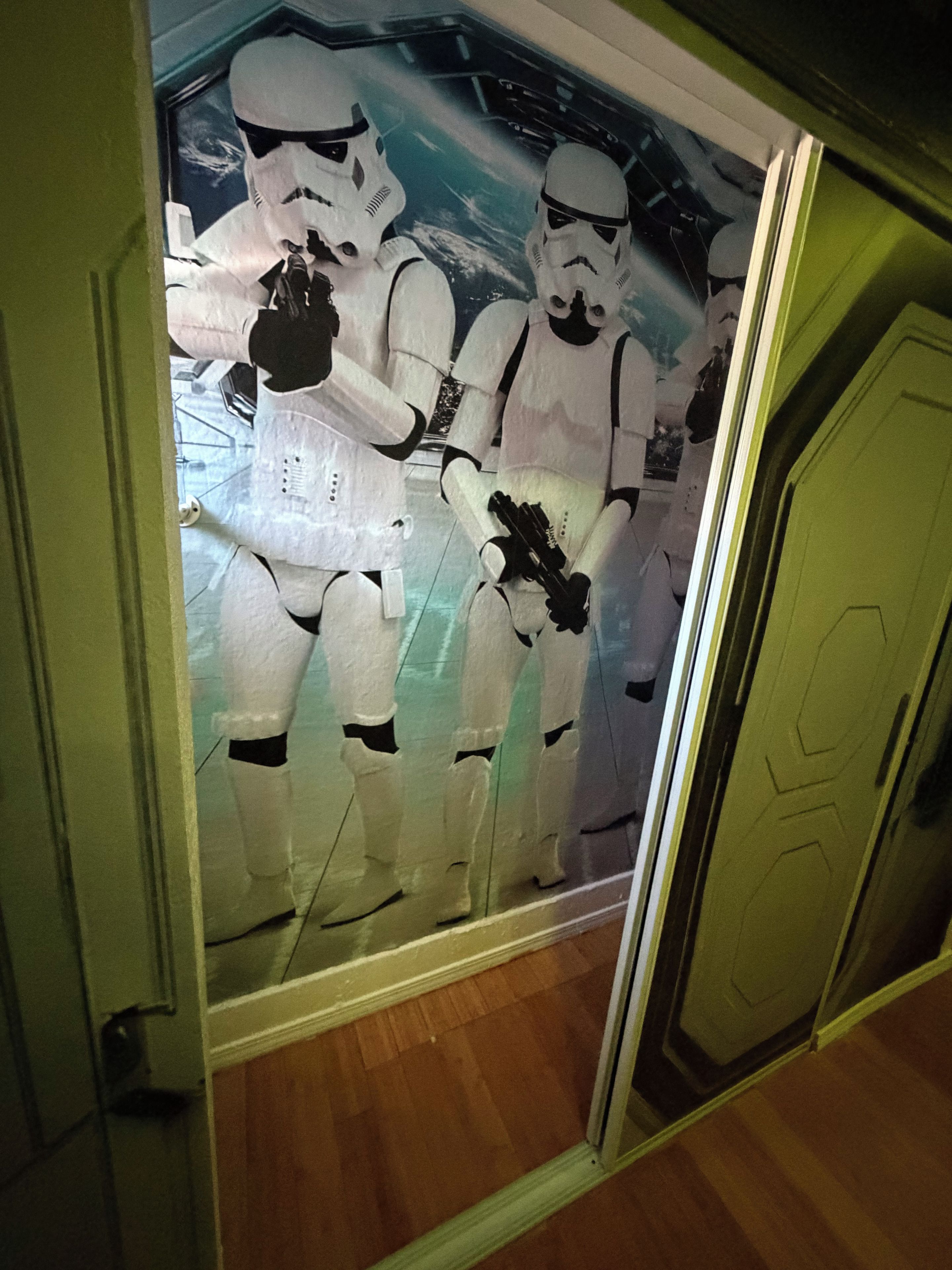 A room with a door leading to a hallway, a wooden floor, and a wall with a poster of stormtroopers.