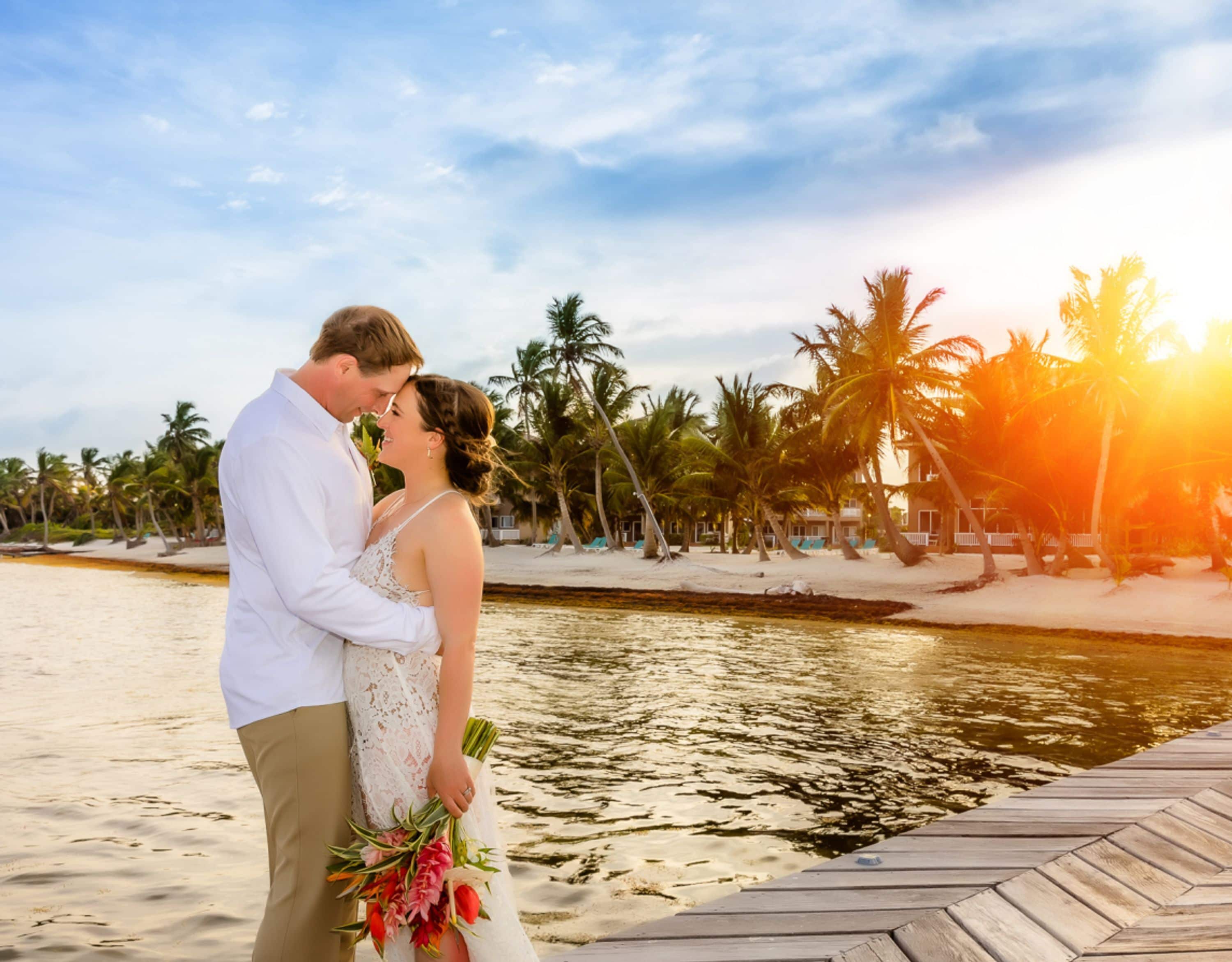A couple standing on a wooden dock by a body of water with palm trees and a beach in the background.