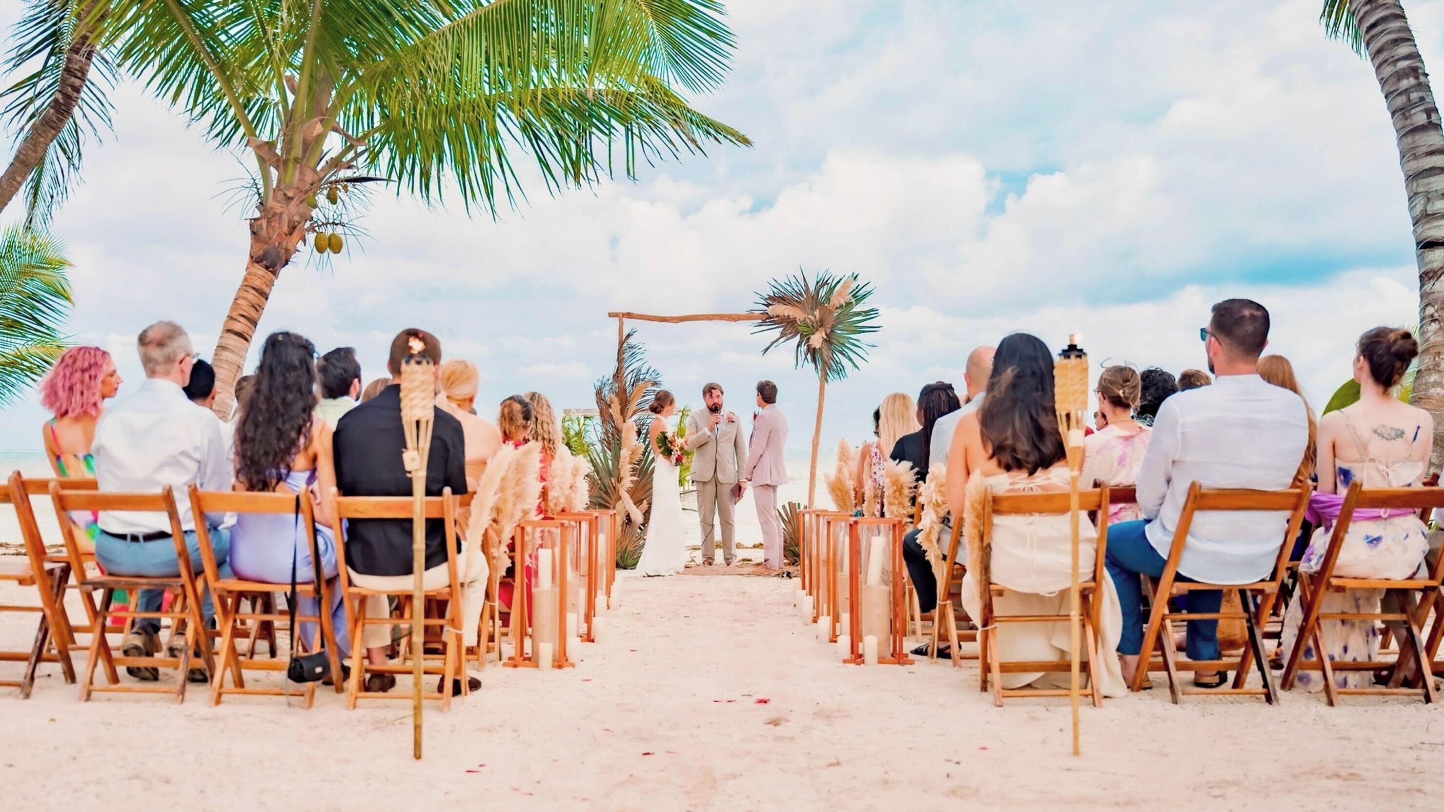 Une cérémonie de mariage sur la plage, les invités étant assis sur des chaises en bois, face à une allée centrale.