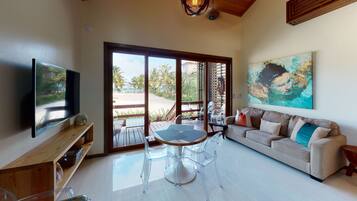 Una sala de estar con televisor, sofá, mesa con sillas y un amplio ventanal con vista a palmeras y una playa.