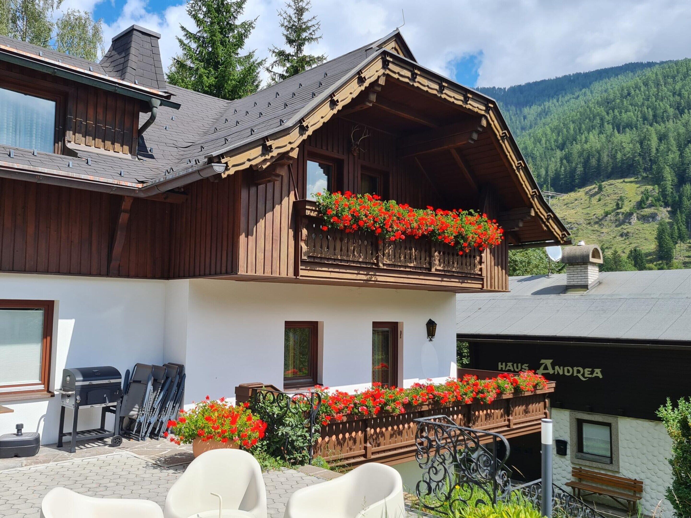 A chalet with a balcony adorned with red flowers, a barbecue area, and a view of the mountains.