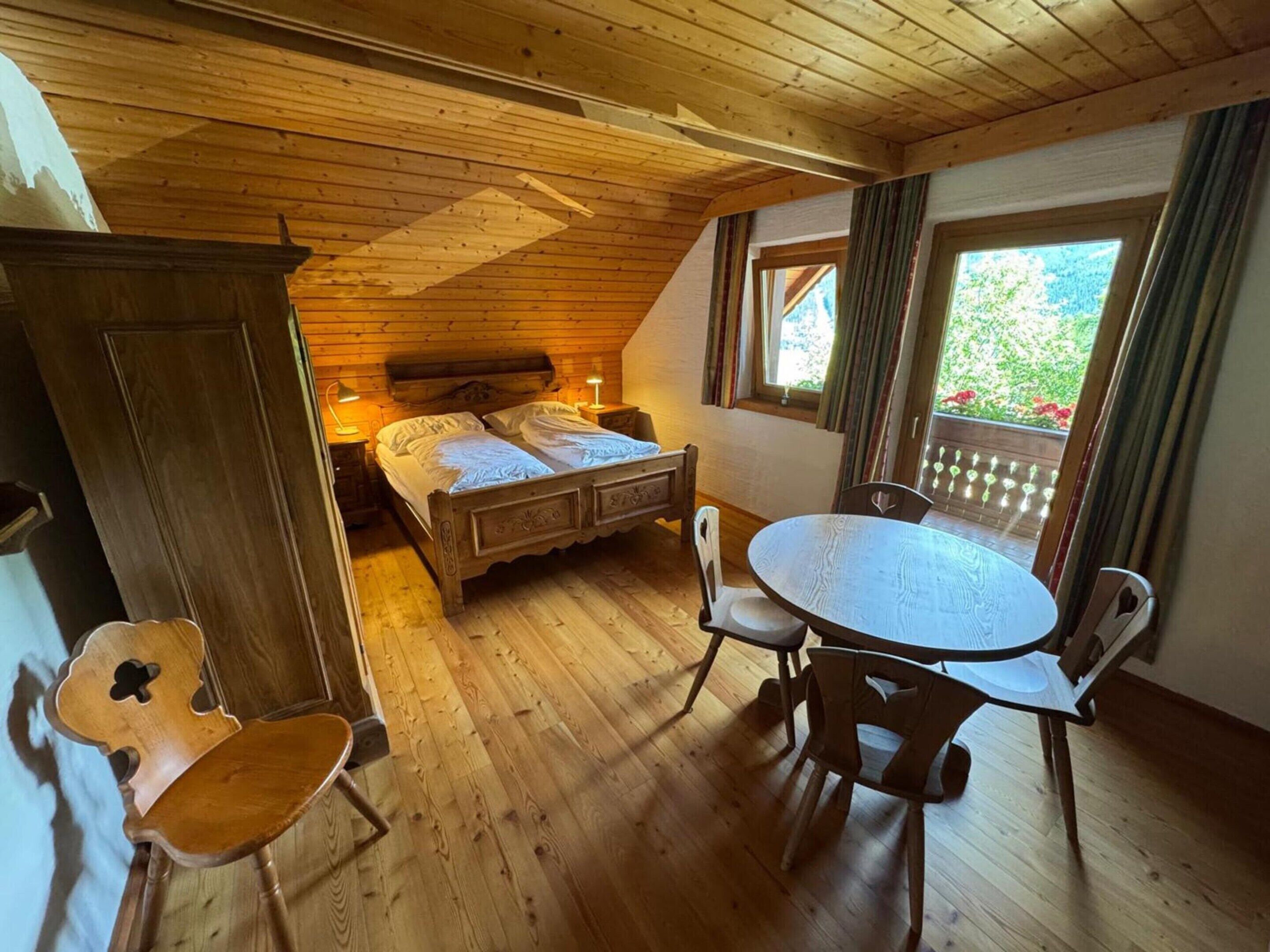 A room with a wooden ceiling, a bed with a headboard, a round table with chairs, and a window with curtains.