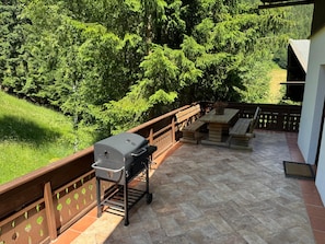 An outdoor patio with a barbecue grill, wooden benches, and a view of a forested area. - Landhaus Finkenhof (Bad Kleinkirchheim)