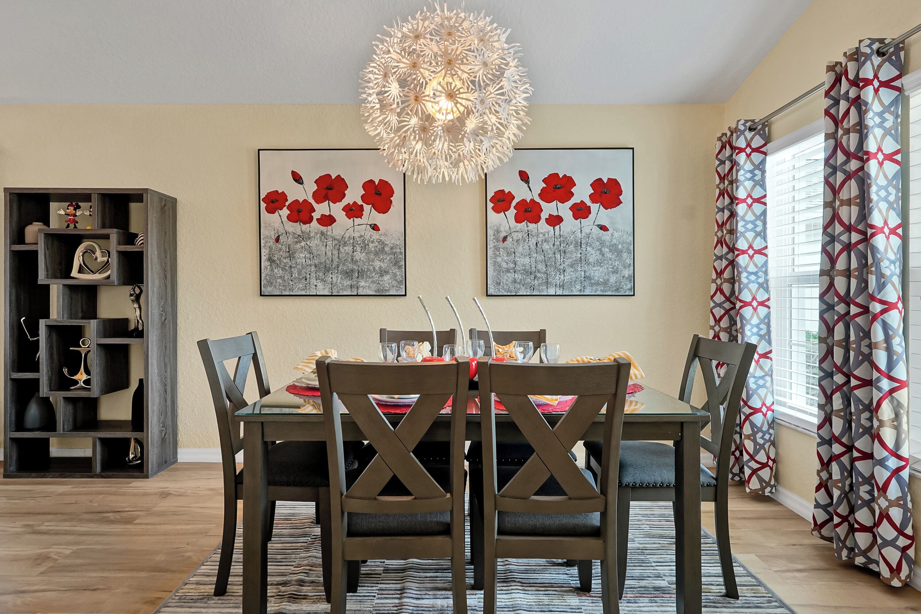 Un coin repas avec une table dressée pour quatre personnes, entourée de chaises dont le dossier présente un motif en croisillons. Deux œuvres d’art encadrées, représentant des fleurs rouges, sont accrochées au-dessus de la table. Un lustre décoratif est suspendu au plafond, et un rideau à motifs est visible sur la droite.