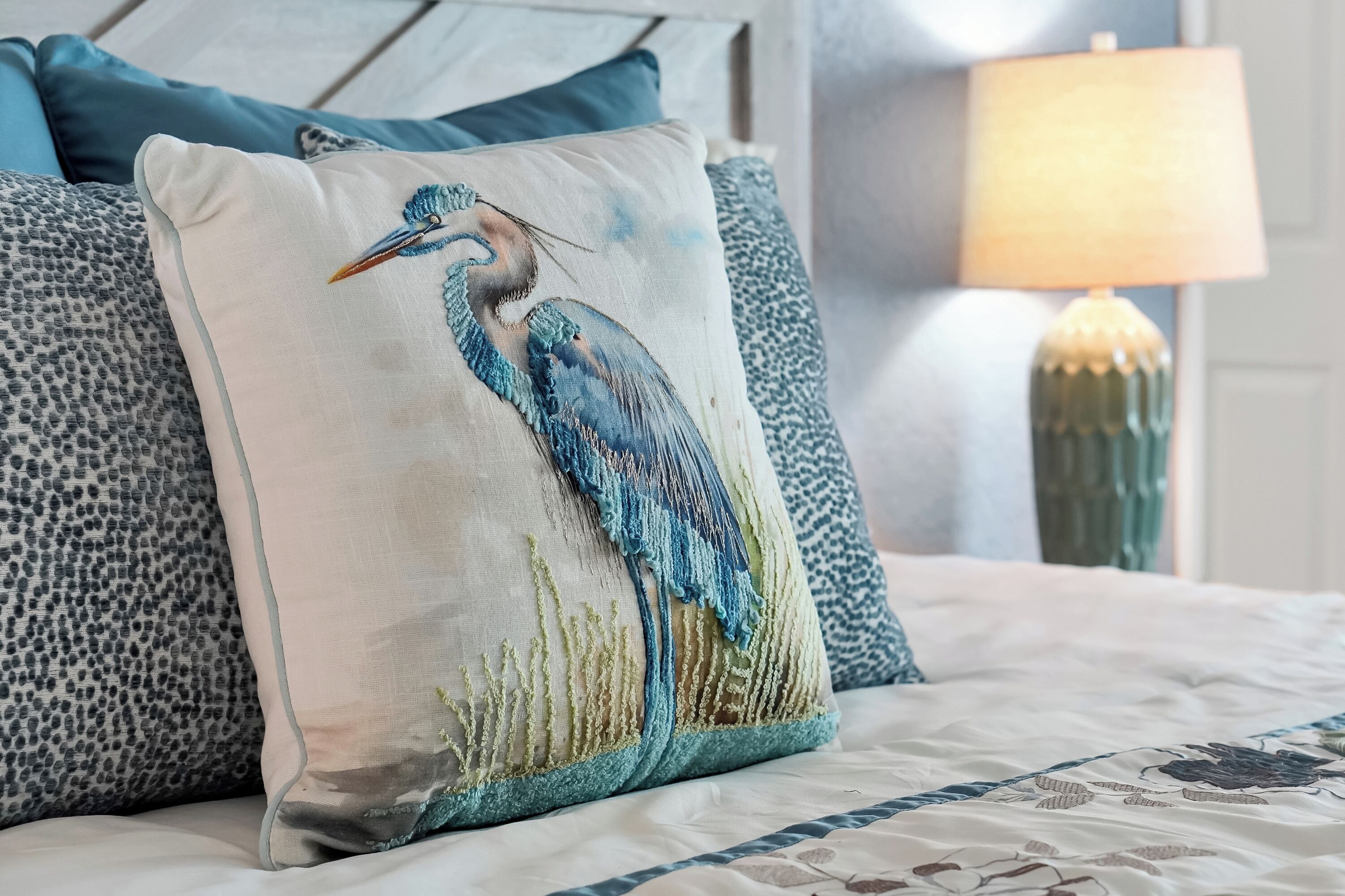 Ein Bett mit einem Vogel-Motiv auf dem Kopfkissen, einem gemusterten Kissen und einer Lampe im Hintergrund.
