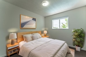 A bedroom with a bed, nightstands, a lamp, a bedside table with books, a window with a view, and a plant.