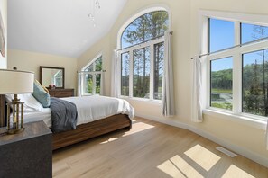 A bedroom with a large bed, a nightstand, a lamp, and multiple windows offering views of a forest and a lake.