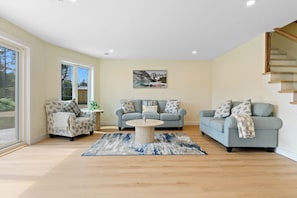 A spacious living room with a large window, a sofa set, a coffee table, and a painting on the wall.