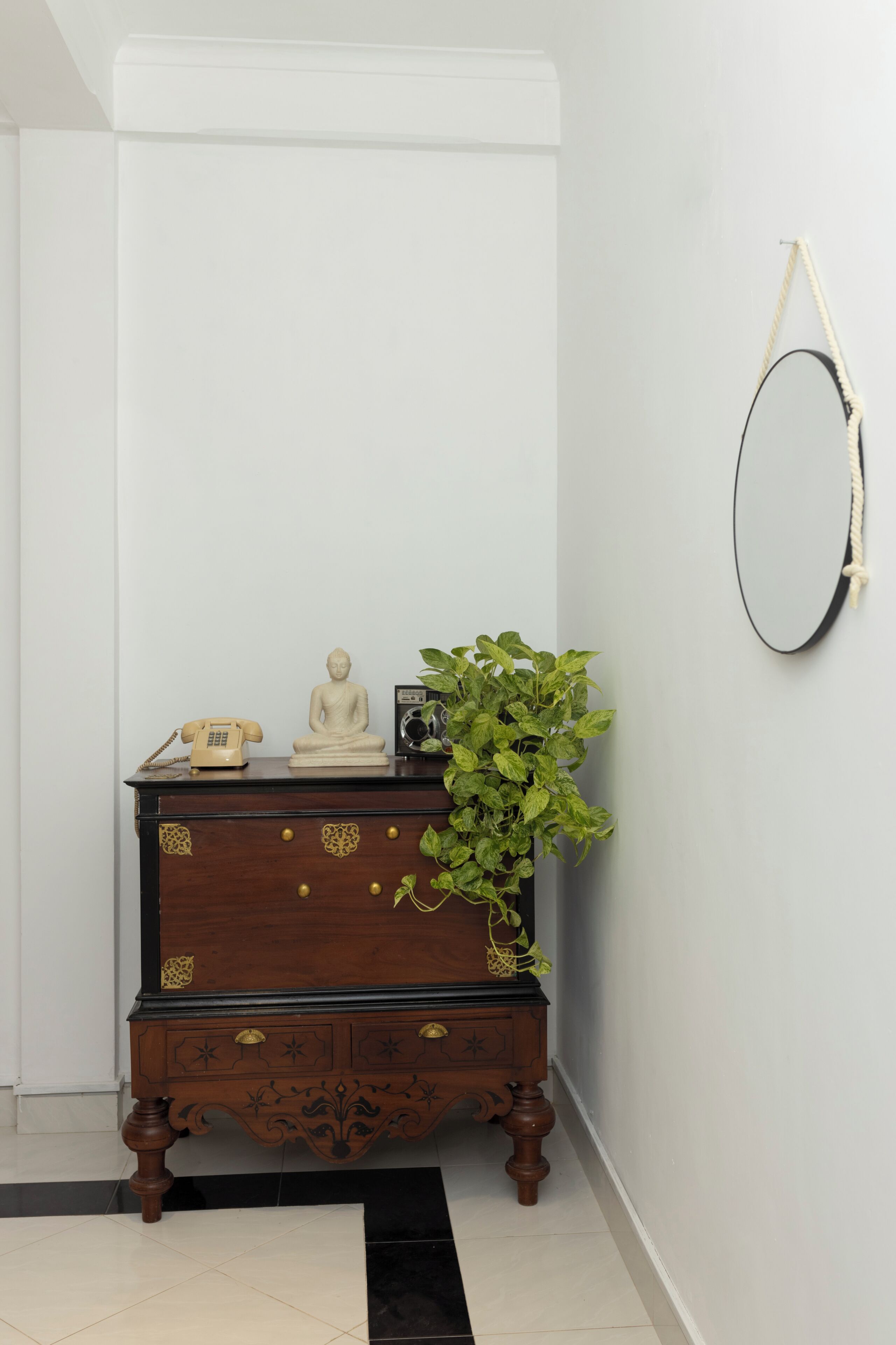 A corner room with a dark wooden dresser, a small Buddha statue, a vintage telephone, and a green plant.