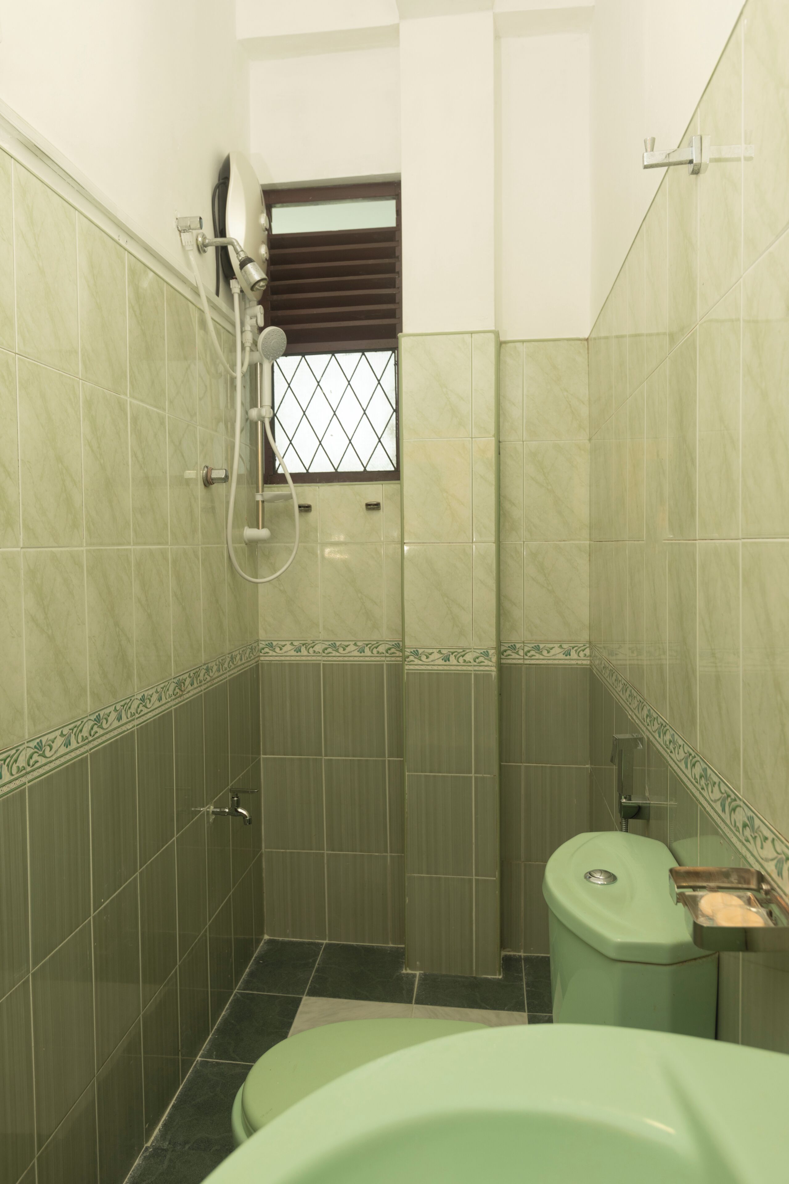 A bathroom with green tiles, a shower, a toilet, and a window with a metal grate.