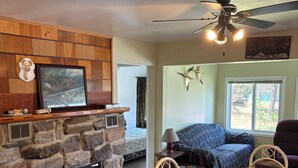 A cozy living room with a stone fireplace, wooden panel walls, a ceiling fan, and a view through a window.