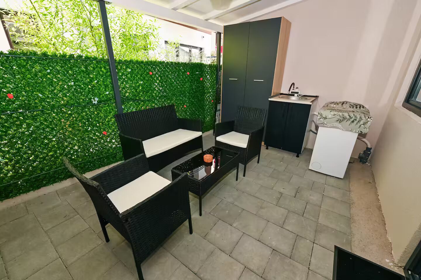 A patio area with a seating arrangement, a small table, a cabinet, and a green hedge.